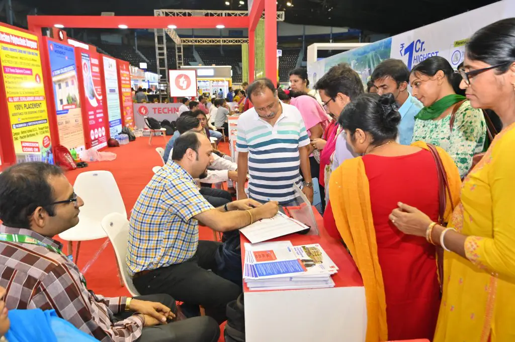 Man in checkered shirt signs a form at a registration desk while a queue of visitors waits at a trade fair booth with colorful banners in the background.