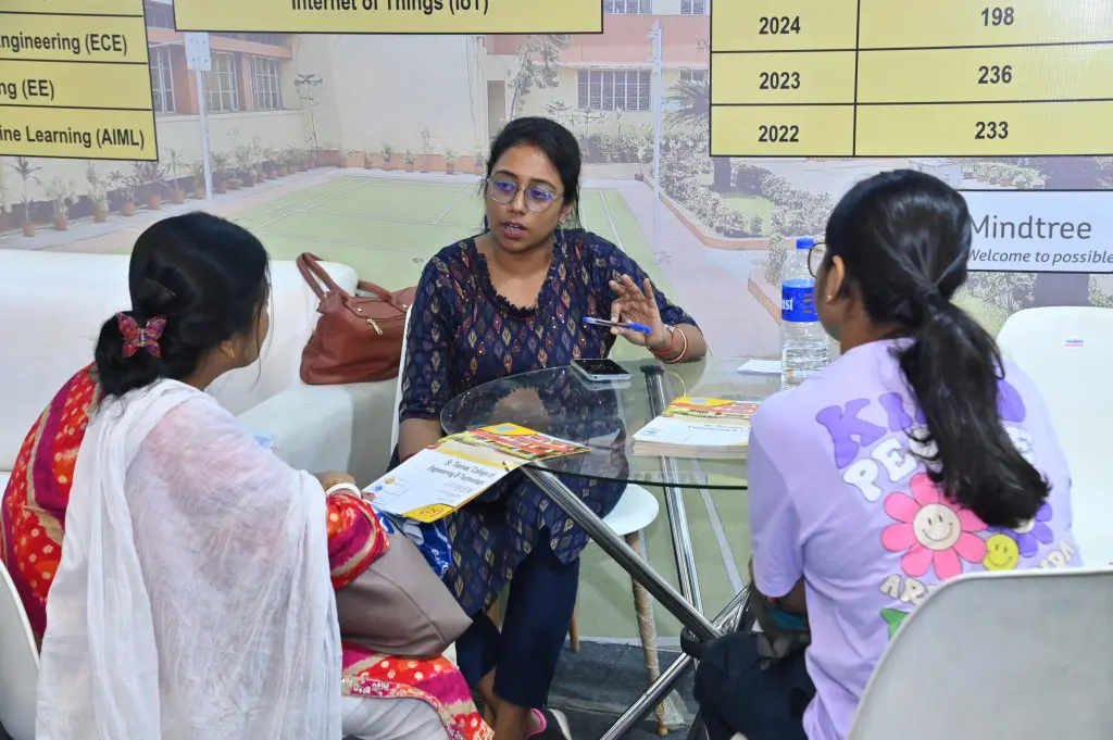 Three women sit around a glass round table in a booth, one woman in a blue patterned top explaining with a hand gesture while two others listen; brochures and a water bottle on the table, backdrop shows campus information.