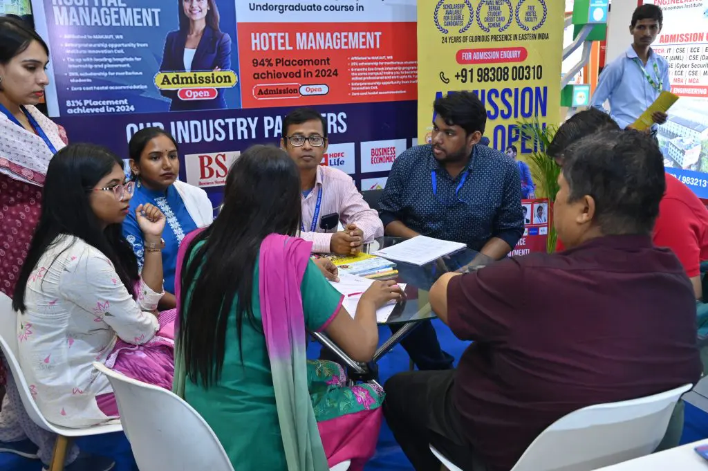 Panel of people at an education fair booth discussing with applicants; posters advertising hotel management and admissions in the background.