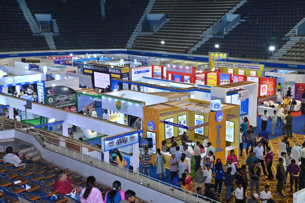 Busy indoor exhibition hall with numerous colorful booths and crowds of people at a trade fair in a stadium-like venue.